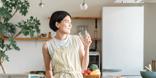 Woman drinking water in a kitchen with fresh whole foods, supporting skin health and nutrition