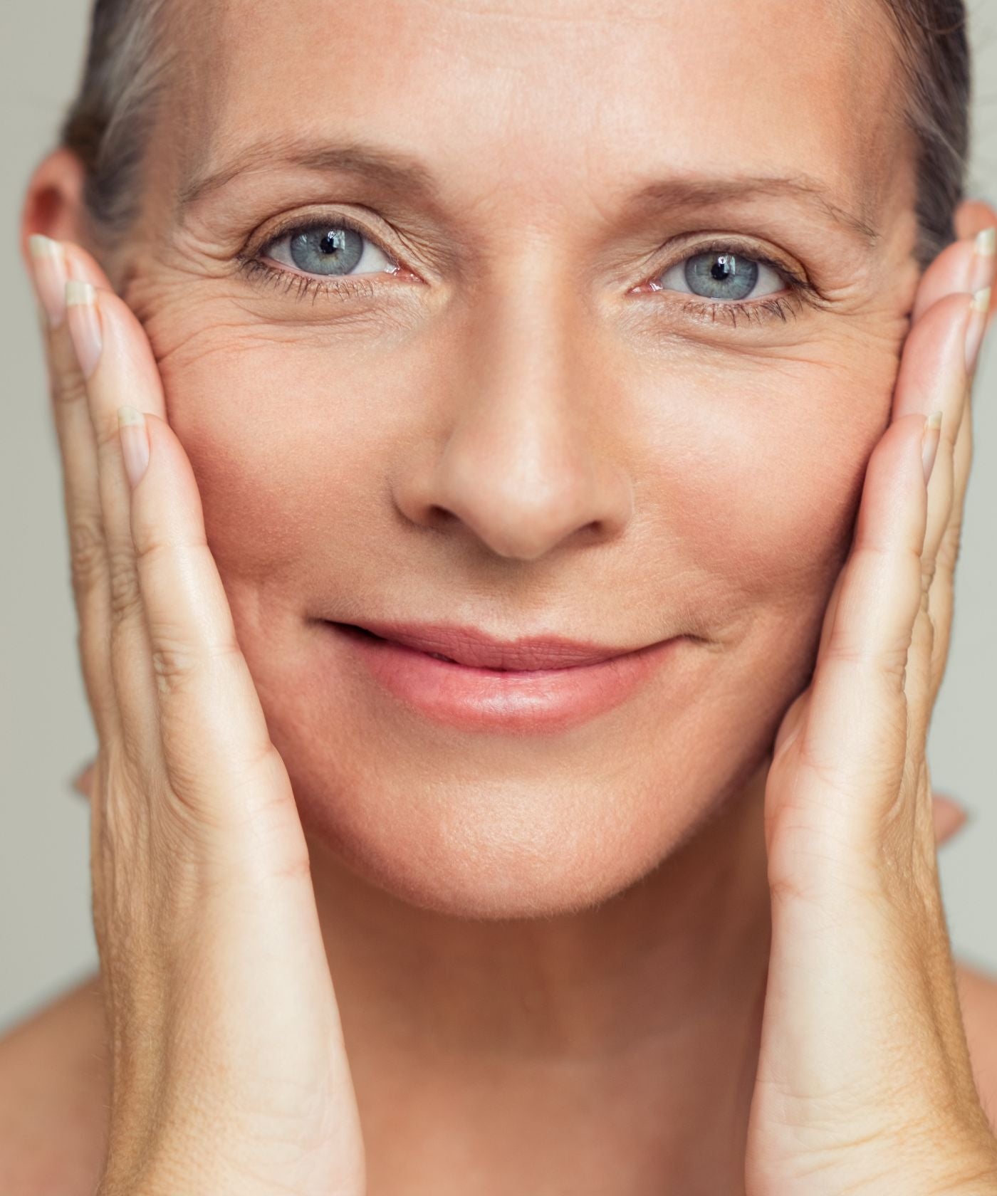 A close-up portrait of a smiling woman with her hands resting on her cheeks, showcasing smooth, radiant skin against a soft neutral background.